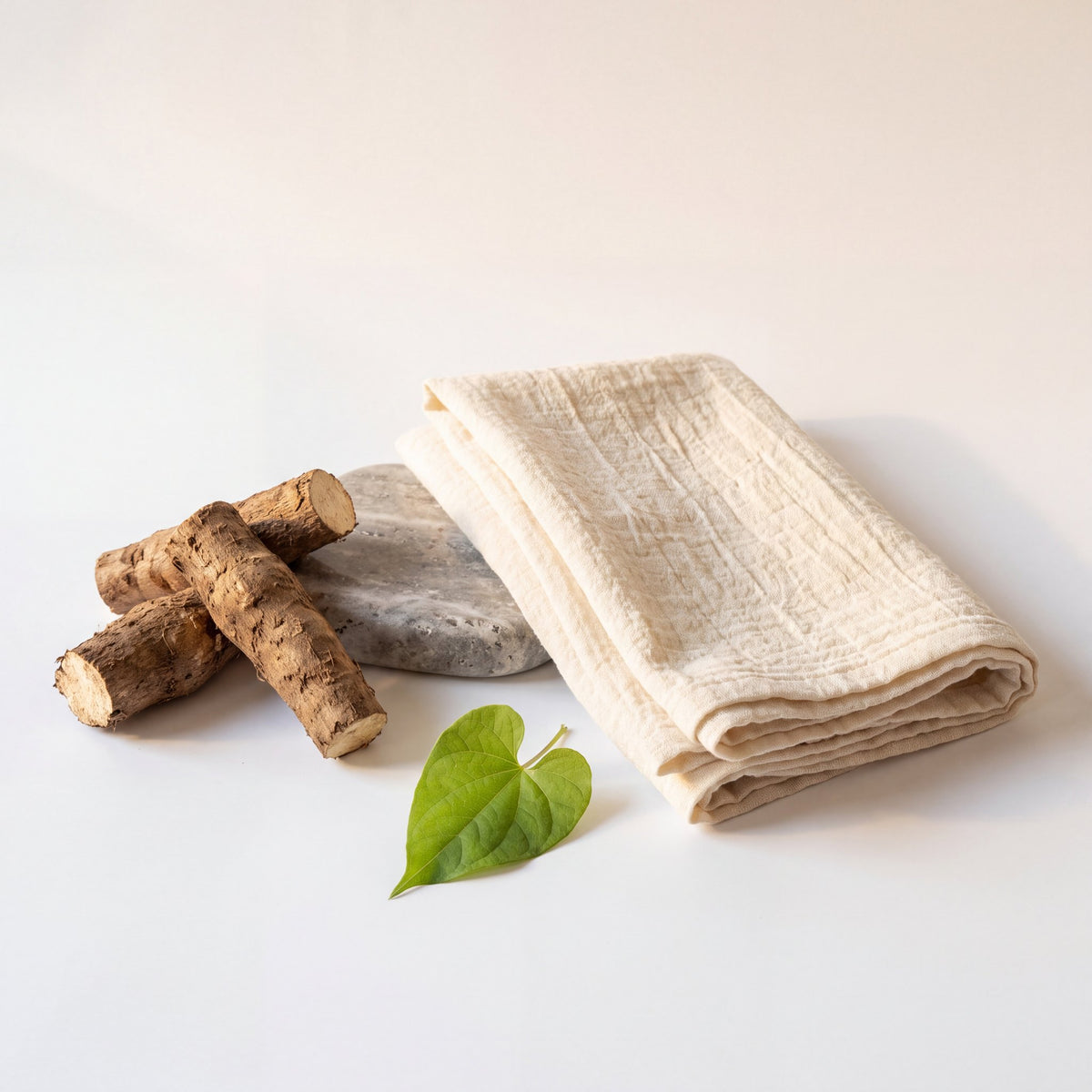 Wild yam roots beside a smooth grey stone, folded ivory linen and a green vine leaf on a white background. Aeshah honest formulation still life